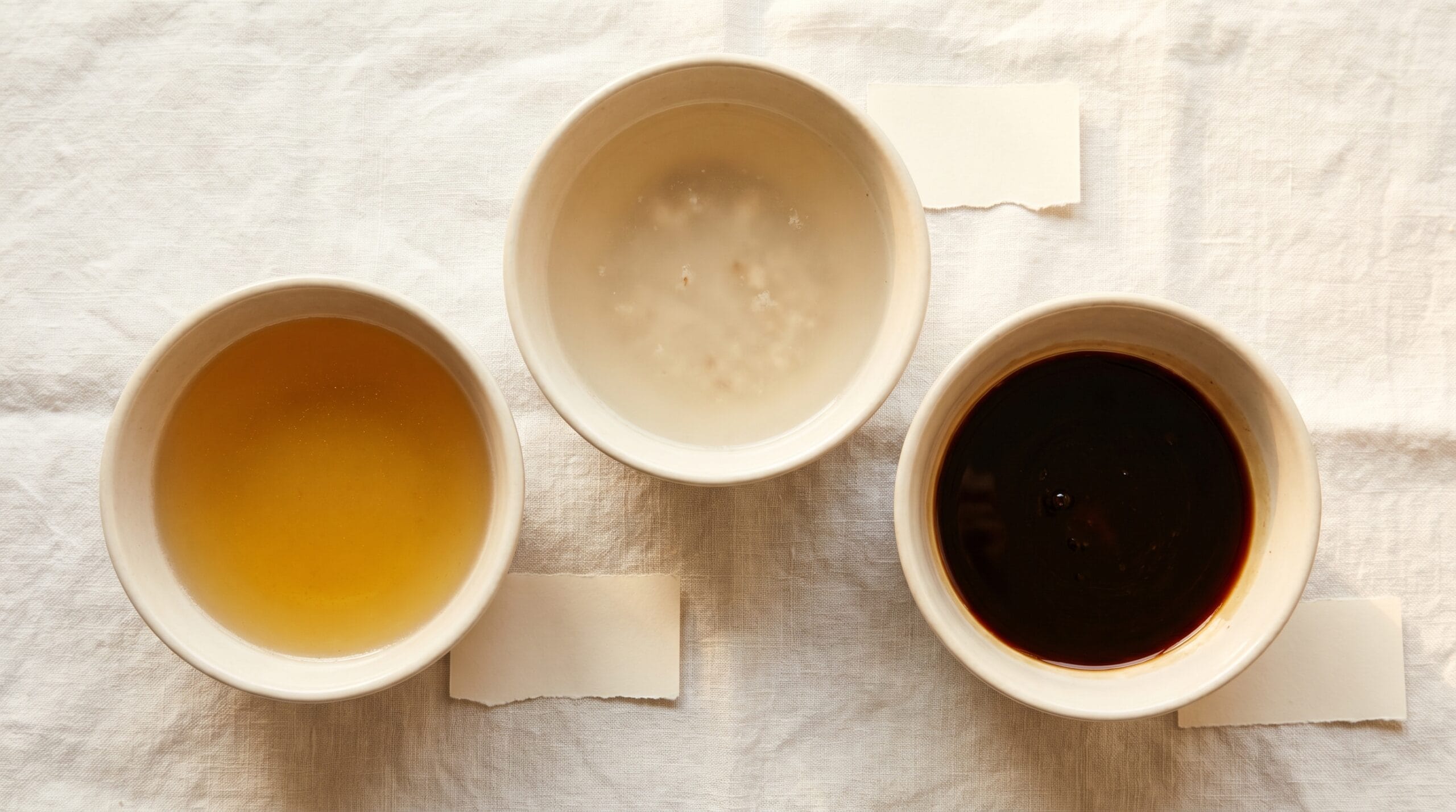 Overhead view of three small ceramic bowls showing clear golden, cloudy pale, and very dark beef broth side by side