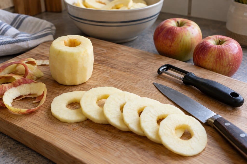 Peeled apple cut into even rings beside a corer hole, peeler, and kitchen knife